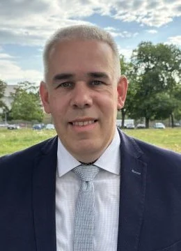 Ein Mann mit schwarzen Anzug und hellblauer Krawatte vor einer Wiese