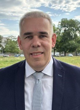 Ein Mann mit schwarzen Anzug und hellblauer Krawatte vor einer Wiese