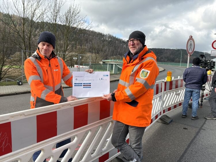 Zwei Männer zeigen den Ablaufplan des Belastungstest an der Elbebrücke in Bad Schandau. Sie stehen auf der Brückenrampe.