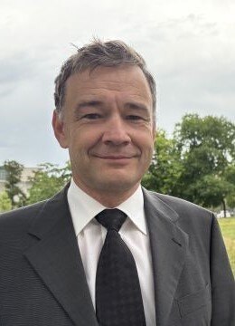 Ein Mann mit einem grauen Anzug und schwarzer Krawatte vor einer Wiese