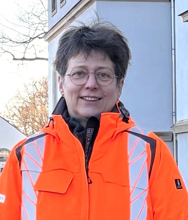 Frau mit Brille und kurzen Haaren mit orangefarbener Jacke vor hellem Gebäude