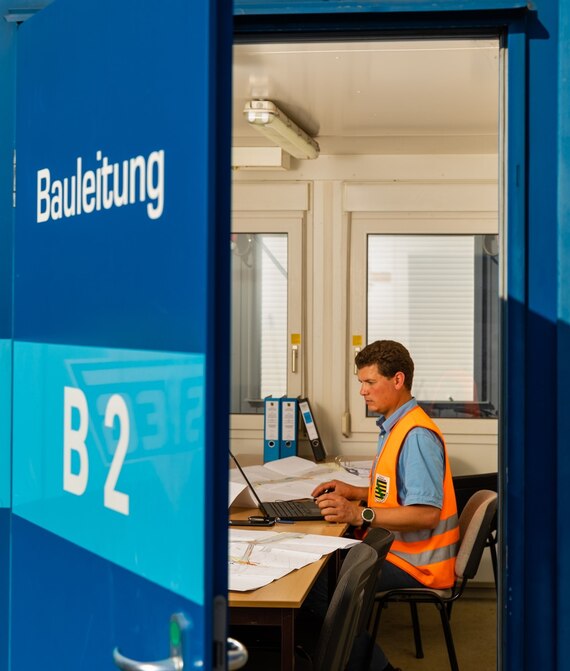 Bauleiter mit oranger Weste in einem Baucontainer mit der Aufschrift Bauleiter B 2 auf der Tür